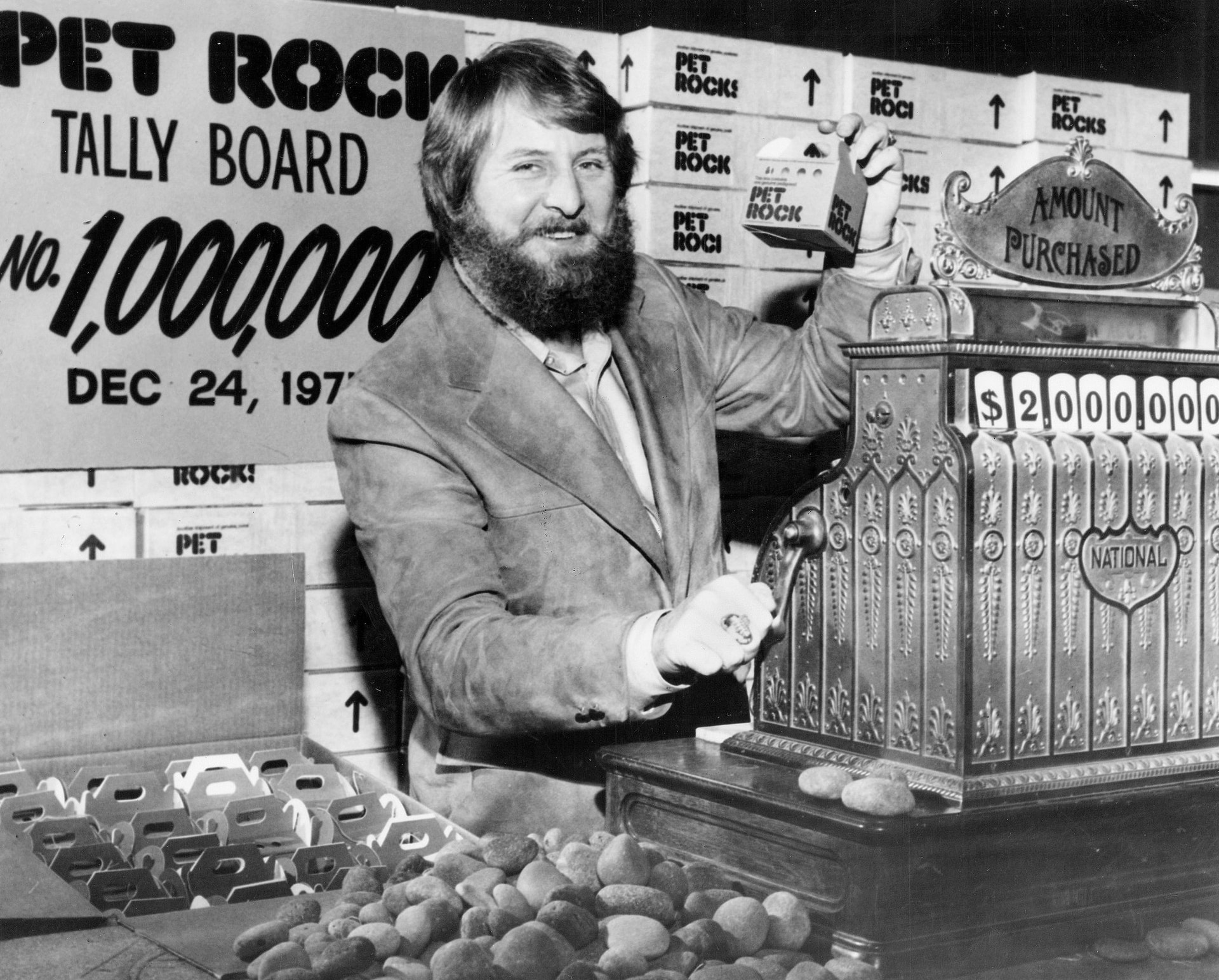Gary Dahl displays a boxed Pet Rock beside a cash register and stacks of Pet Rock packages.